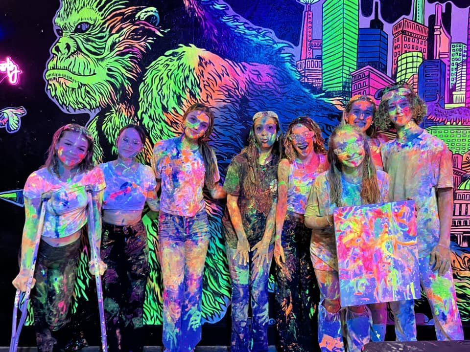 Teenagers covered in neon paint posing before a glowing graffiti wall.