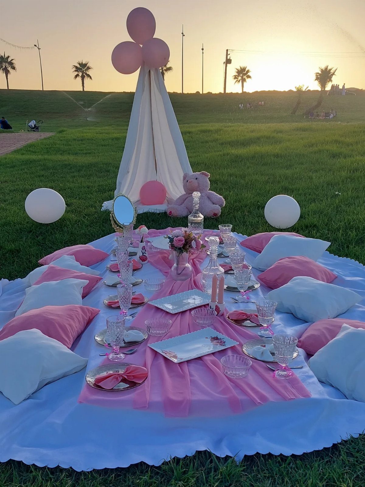 Pink and white picnic setup with cushions and a teepee on a grassy field