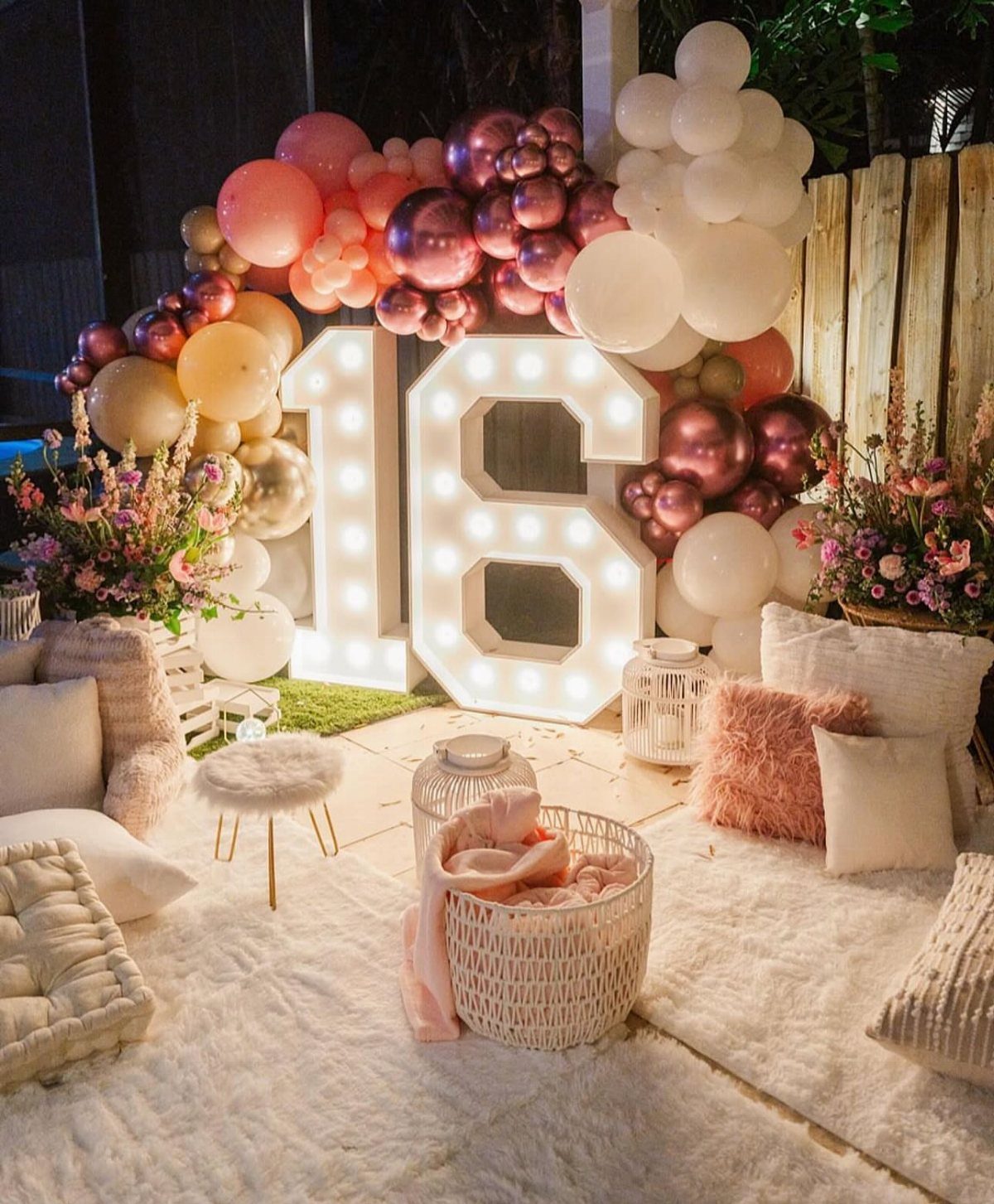 Large illuminated number 16 surrounded by pink and gold balloons and plush pillows