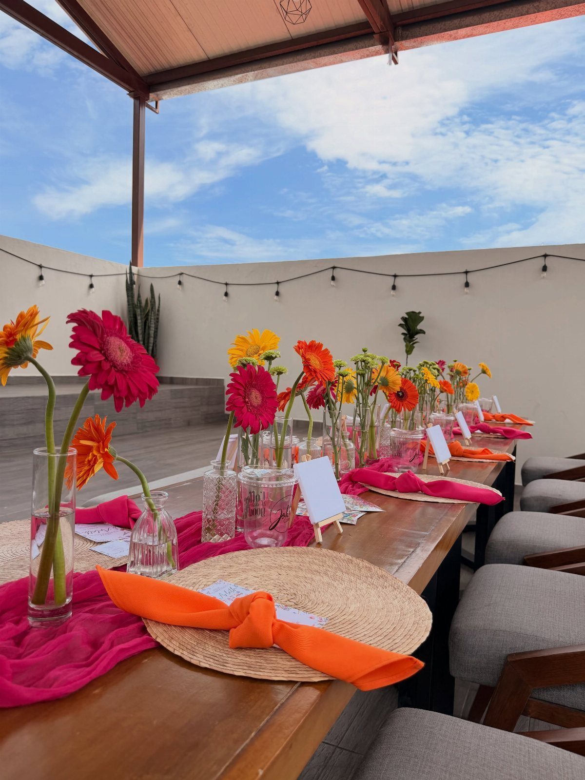 Festive rooftop table setting with pink runners, orange napkins, and colorful daisies.