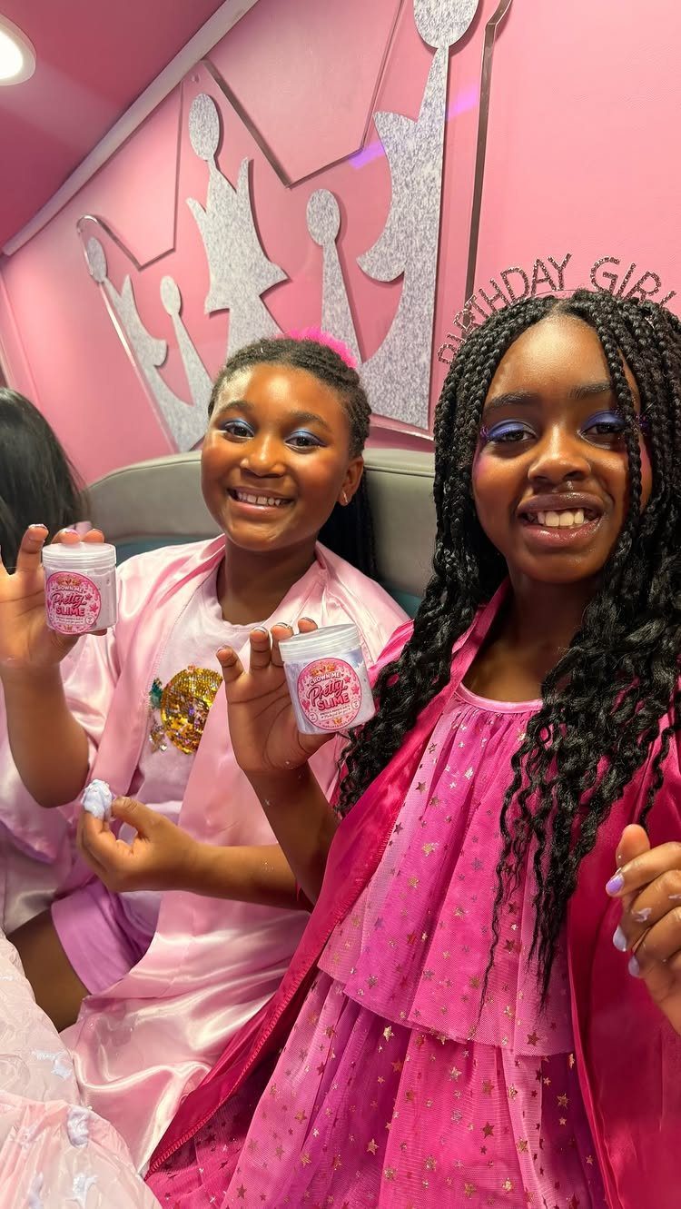 Two girls in pink robes smiling inside a pink party bus with crown decor.