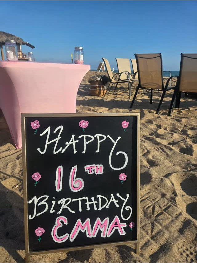 Birthday chalkboard sign on a sandy beach with pink table and chairs.