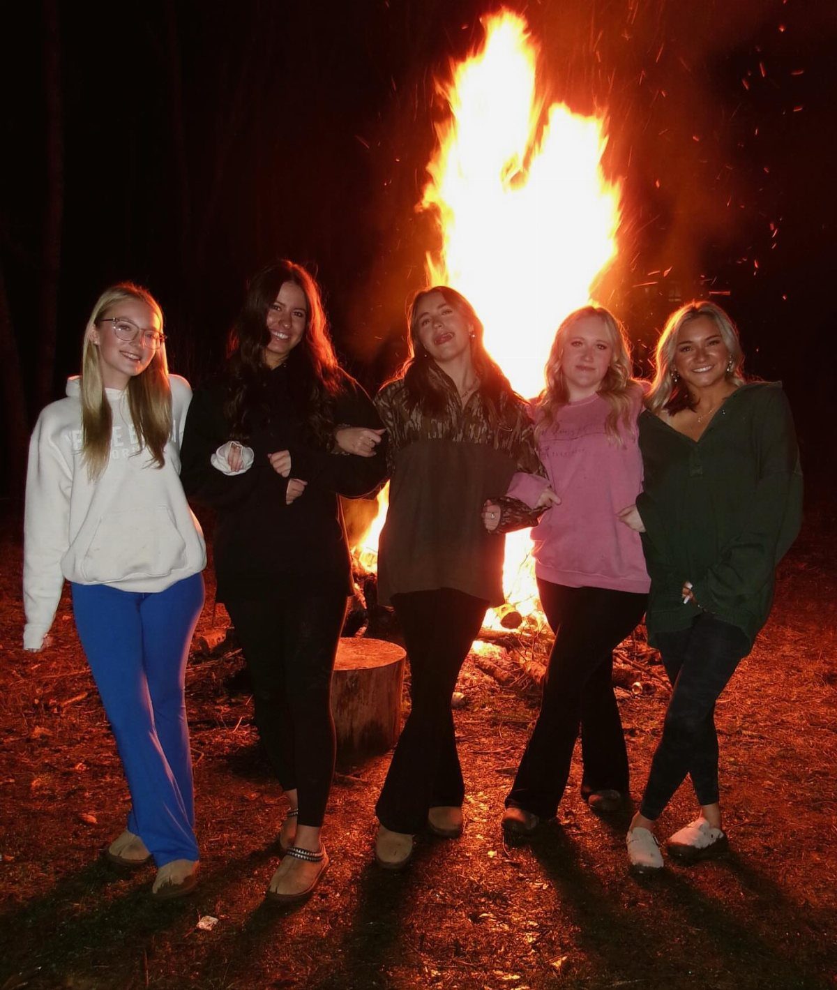 Five teenage girls posing in front of a large glowing bonfire at night.