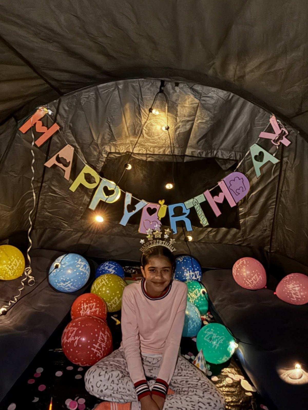 Girl wearing a crown sitting inside a decorated camping tent with birthday balloons.