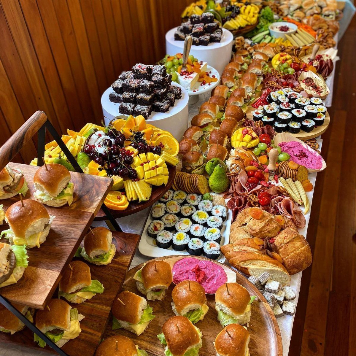 Abundant party food spread with mini burgers, sushi, fruit, and desserts on a table.