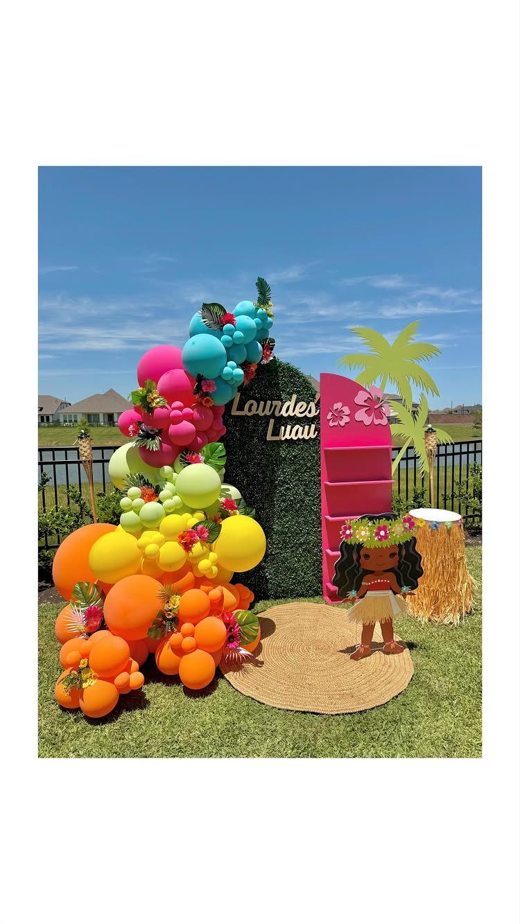 Tropical luau party setup with colorful balloon arch, palm tree, and hula girl.