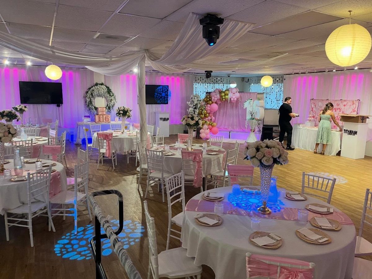 Decorated event hall with white tables, pink lighting, and floral arrangements for a party.