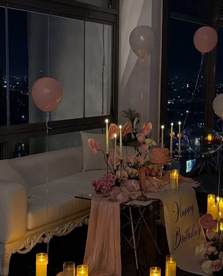 Birthday party setup with pink balloons, candles, and cake in a city apartment.