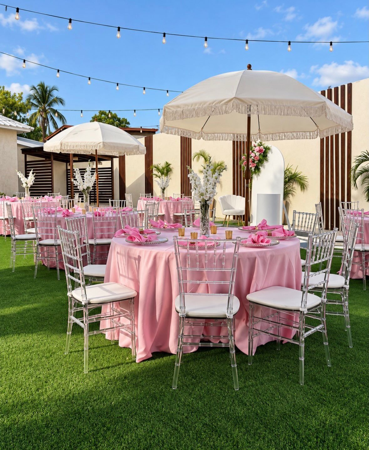 Pink decorated tables and silver chairs on a lawn under white umbrellas.
