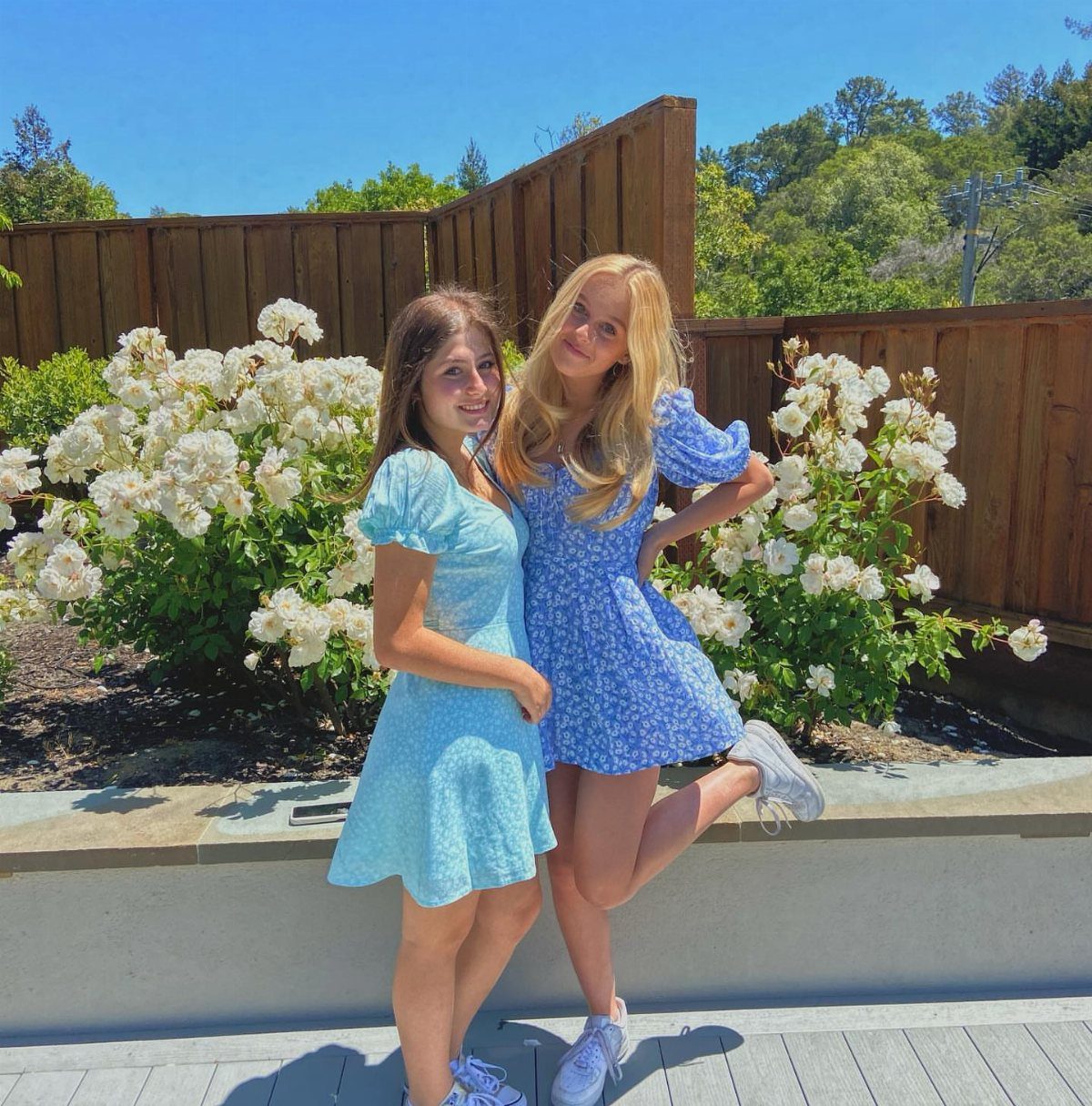 Two teenage girls in blue floral dresses posing together by white rose bushes.