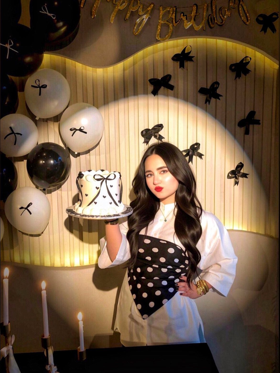 Young woman in polka-dot dress holding a white birthday cake with black bows.