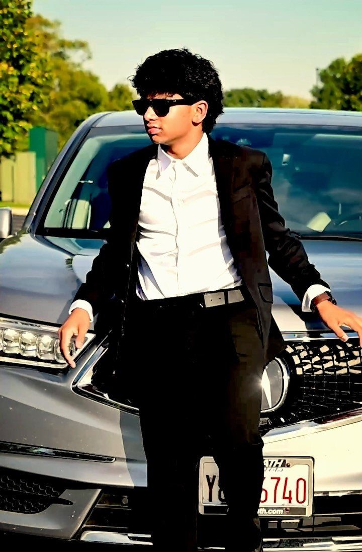 Young man in a black suit and sunglasses posing before a silver car.