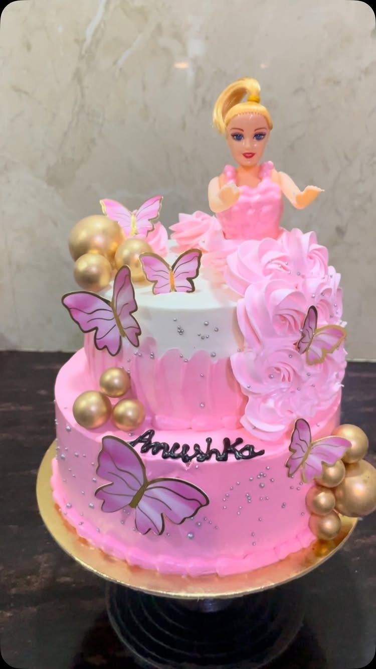 Two-tier pink Barbie cake decorated with frosting roses, purple butterflies, and gold spheres.
