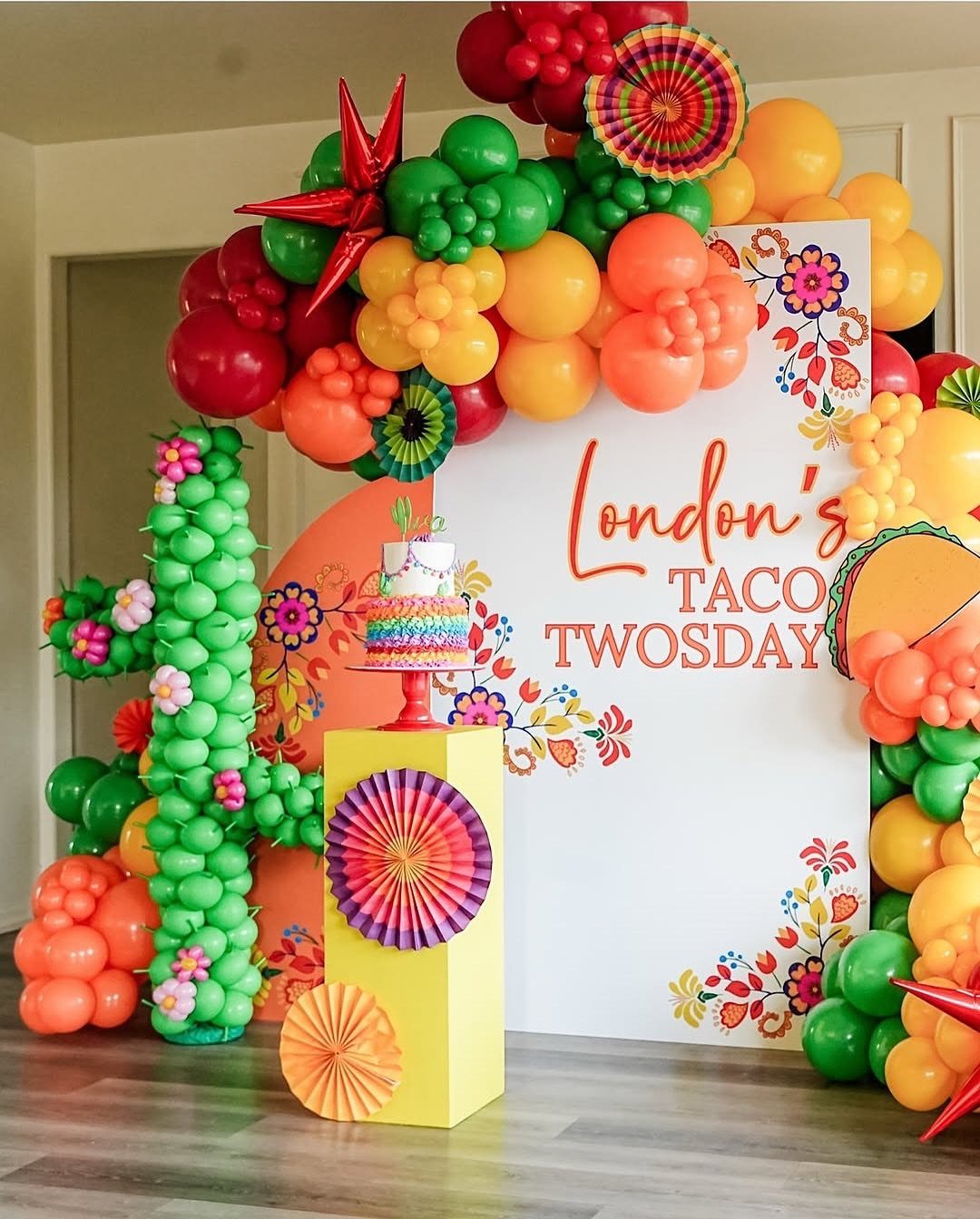 Colorful taco themed party decorations with balloons, a cake, and a custom backdrop.