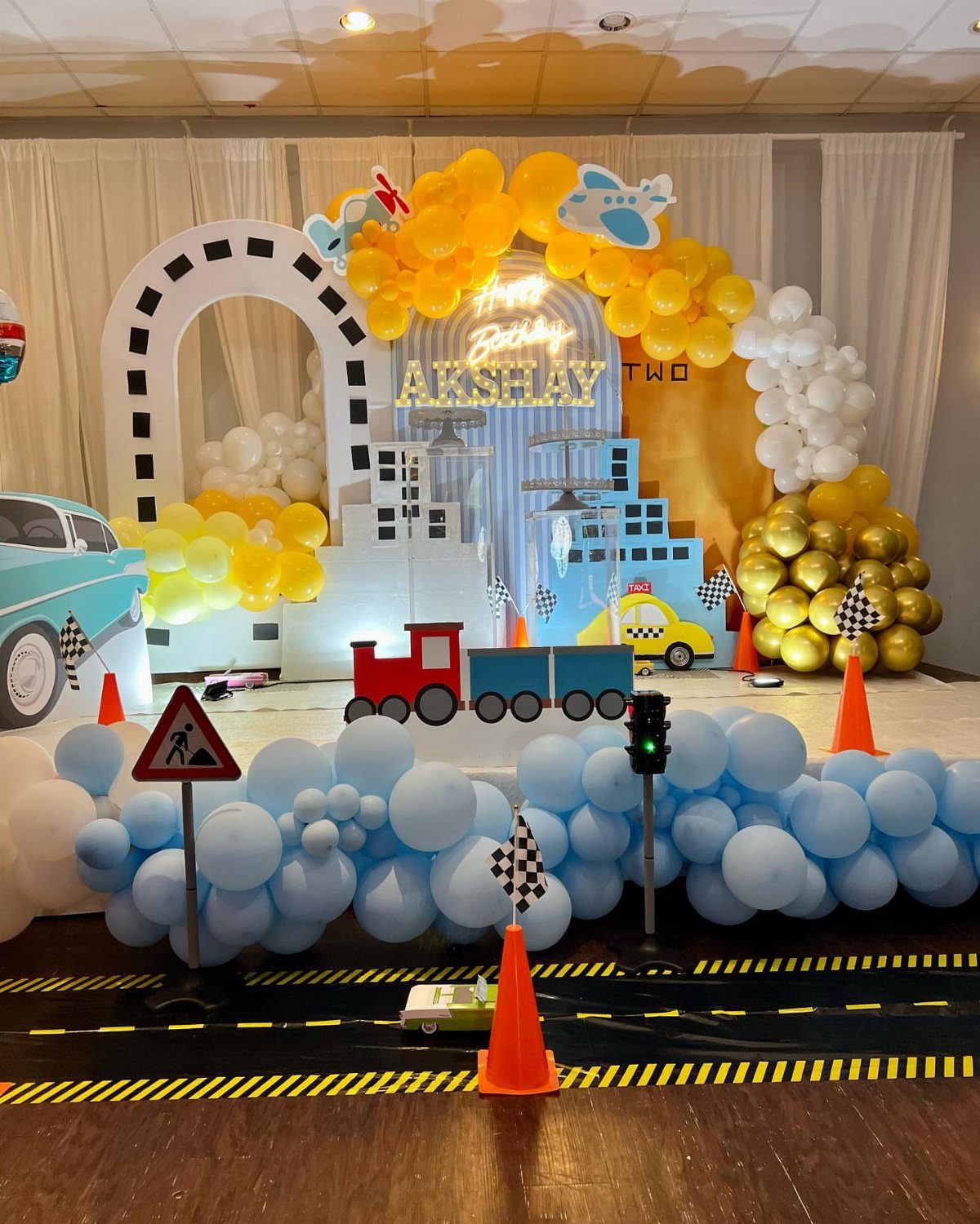 Transportation themed birthday party setup with cars, trains, balloons, and a city backdrop.