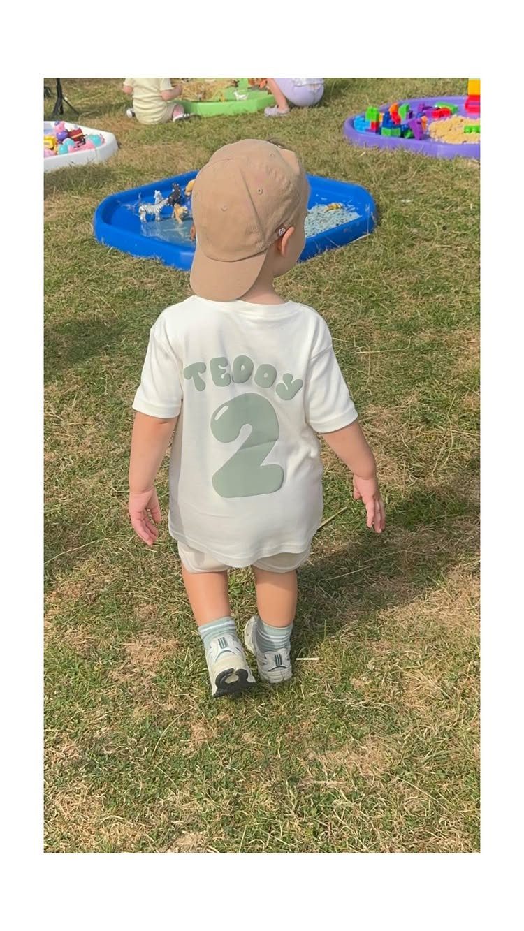 Toddler in a birthday shirt standing before colorful water play sensory bins outdoors.