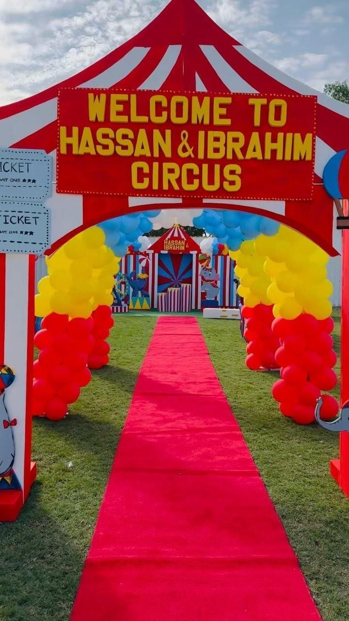 Circus tent entrance with red carpet and colorful balloon arches on green grass.