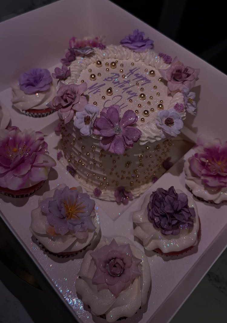 White birthday cake and cupcakes decorated with purple sugar flowers and gold pearls