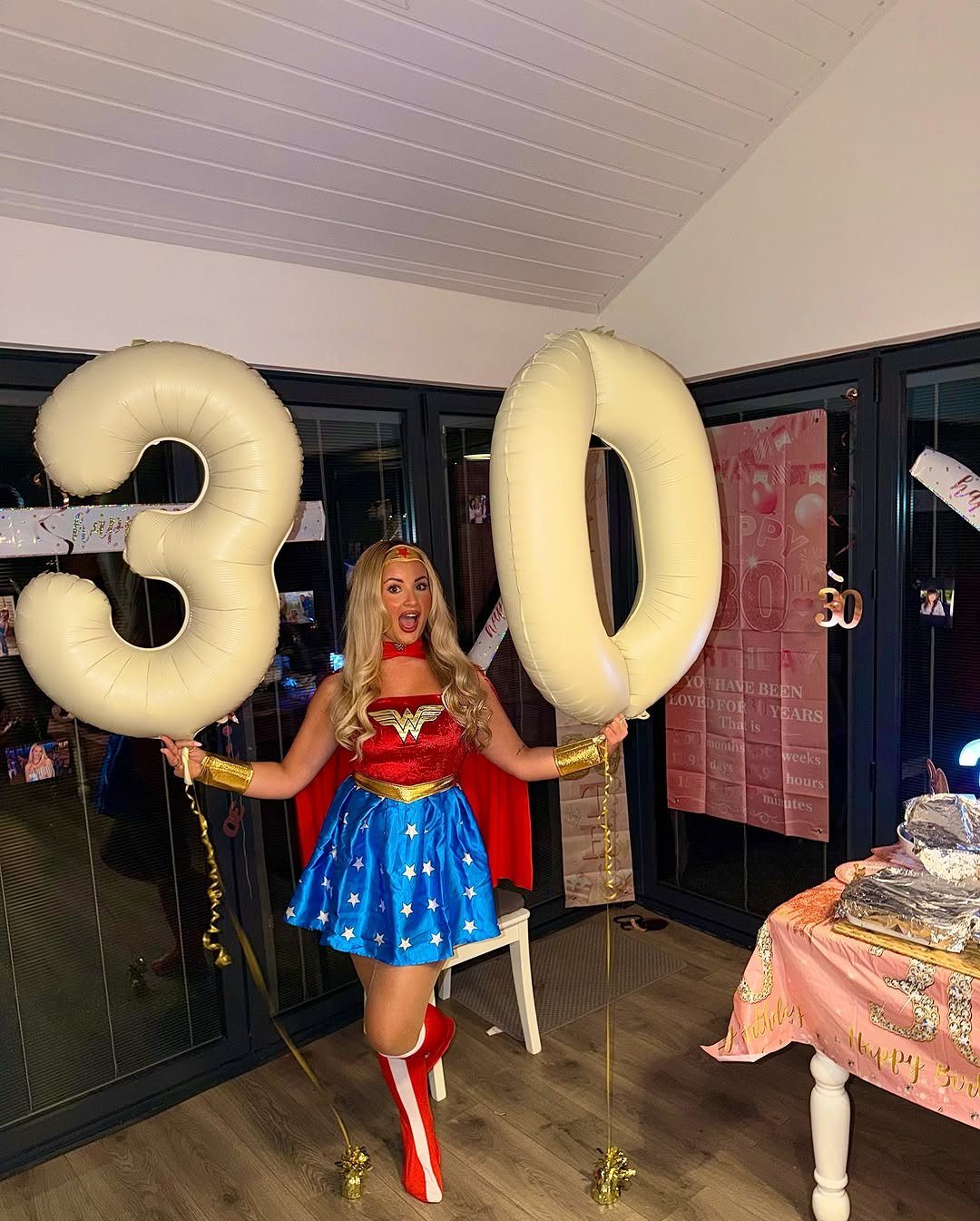 Woman in Wonder Woman costume posing with large number 30 balloons indoors.