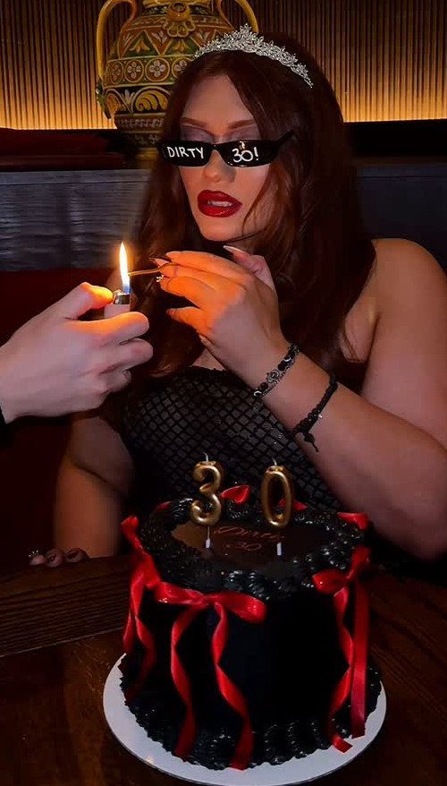 Woman in tiara and Dirty 30 sunglasses lighting candles on a black cake.