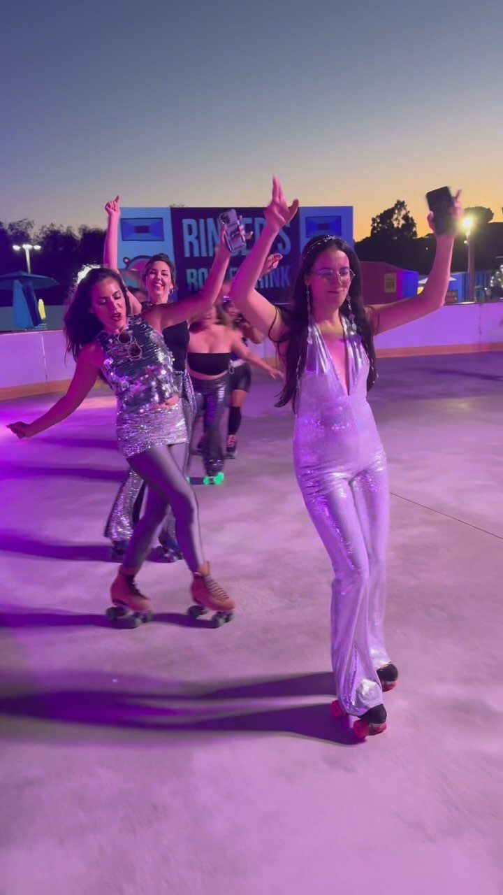Women in silver sequin outfits roller skating on a purple lit outdoor rink.