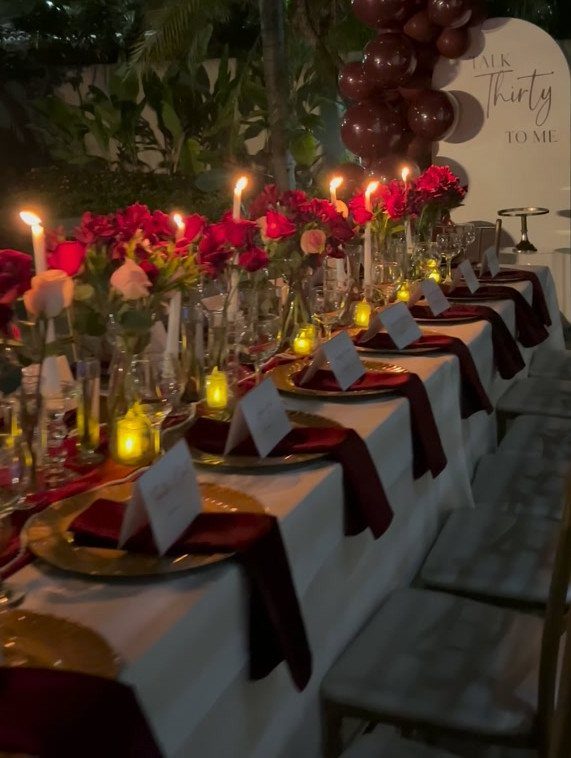 Elegant table setting with gold plates, burgundy napkins, red roses, and lit candles.