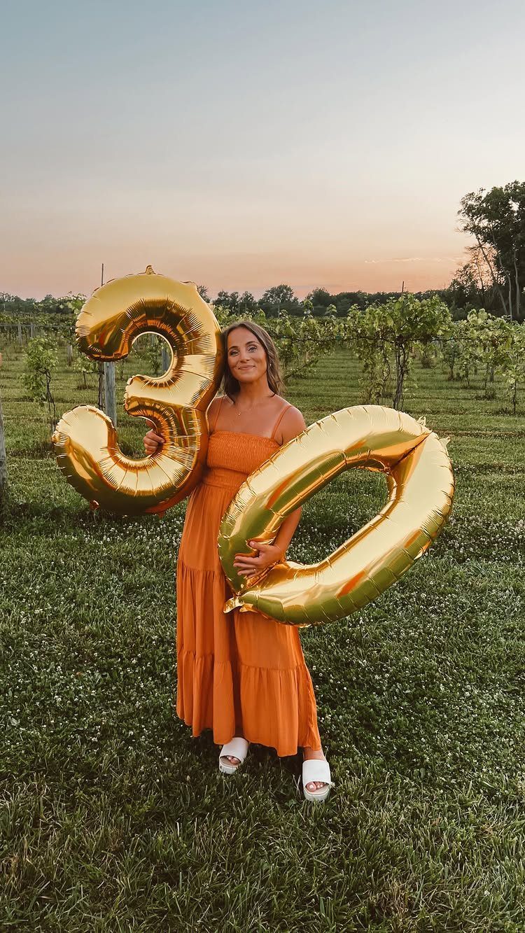 Woman in orange dress holding gold 30 balloons in a vineyard setting