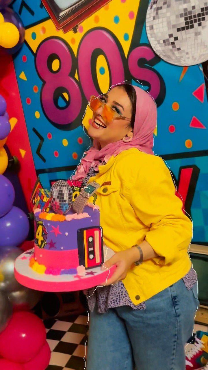 Woman in 80s attire holding a themed cake against a colorful retro backdrop.