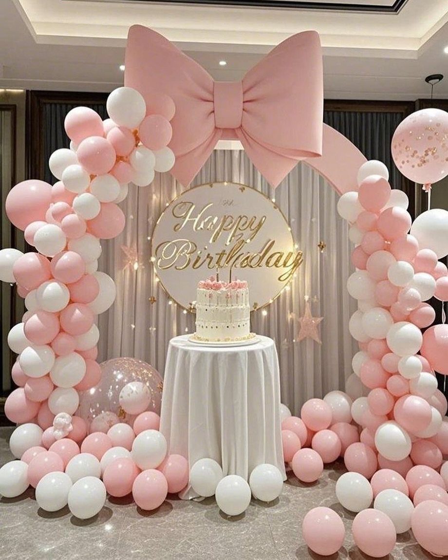 Birthday party setup with pink and white balloon arch, large bow, and cake.
