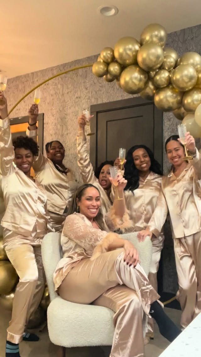 Six women in champagne satin pajamas posing with champagne glasses and gold balloons.