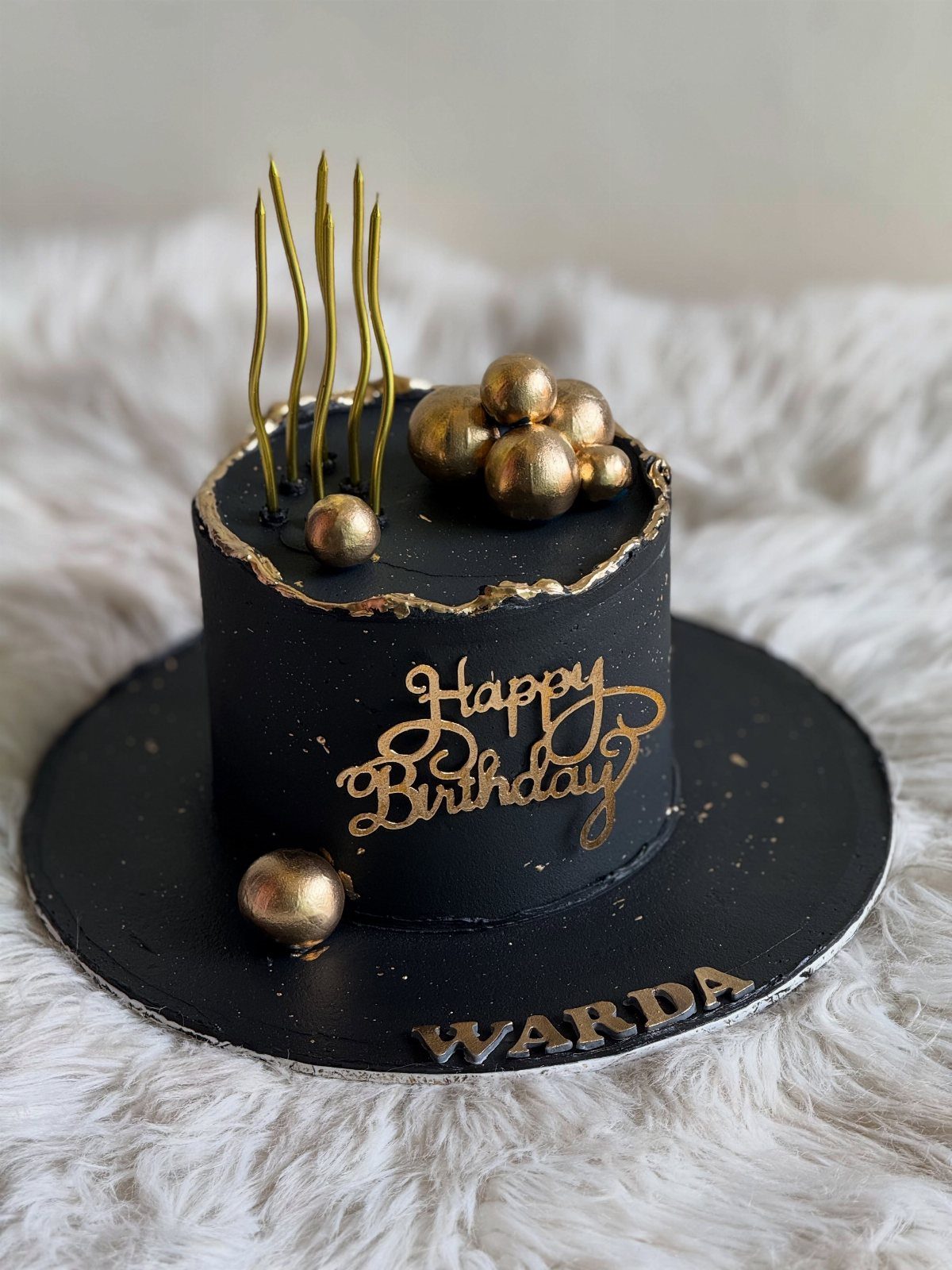 Black birthday cake with gold spheres, gold candles, and gold Happy Birthday lettering.