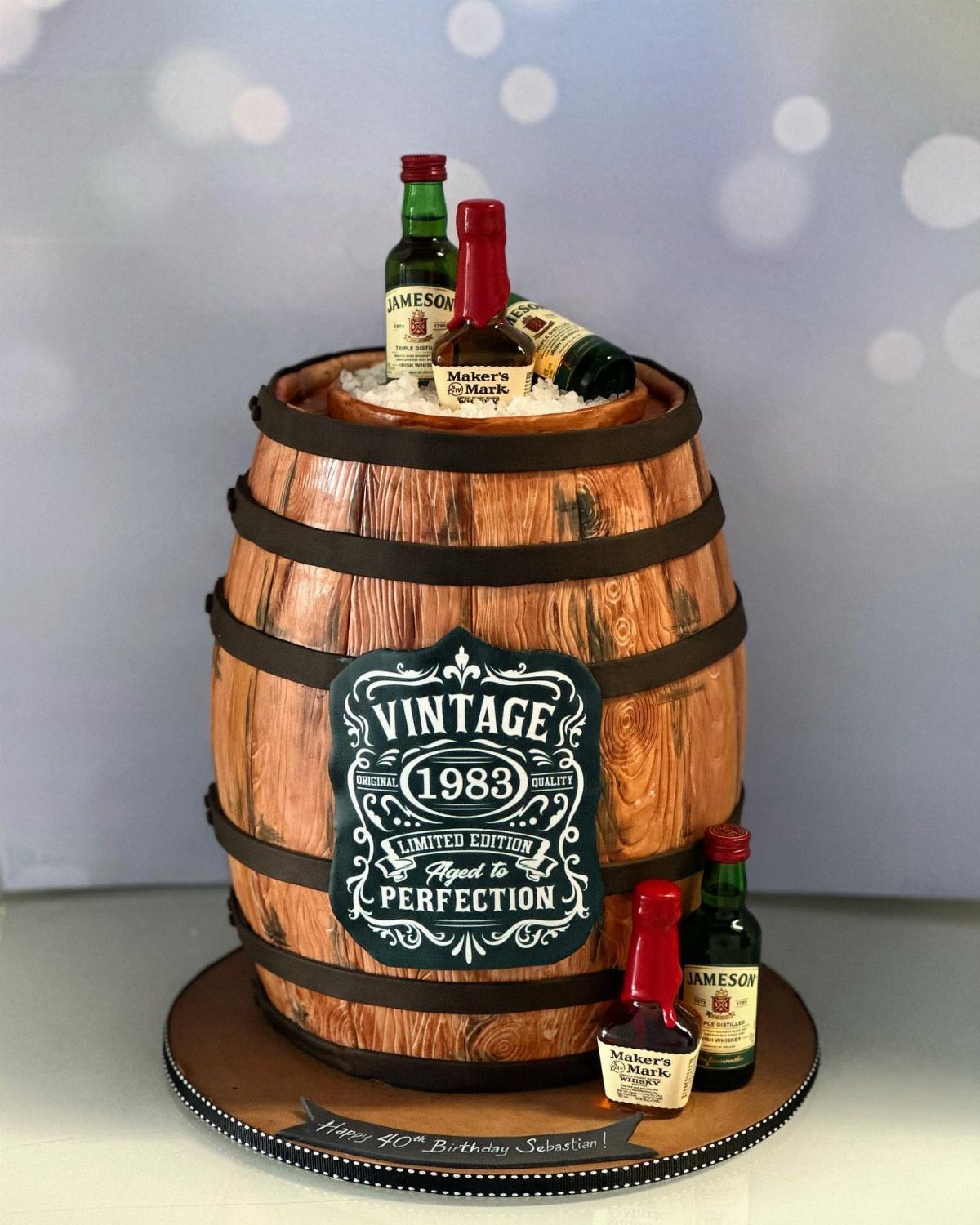 Whiskey barrel shaped cake with vintage 1983 label and miniature whiskey bottles.