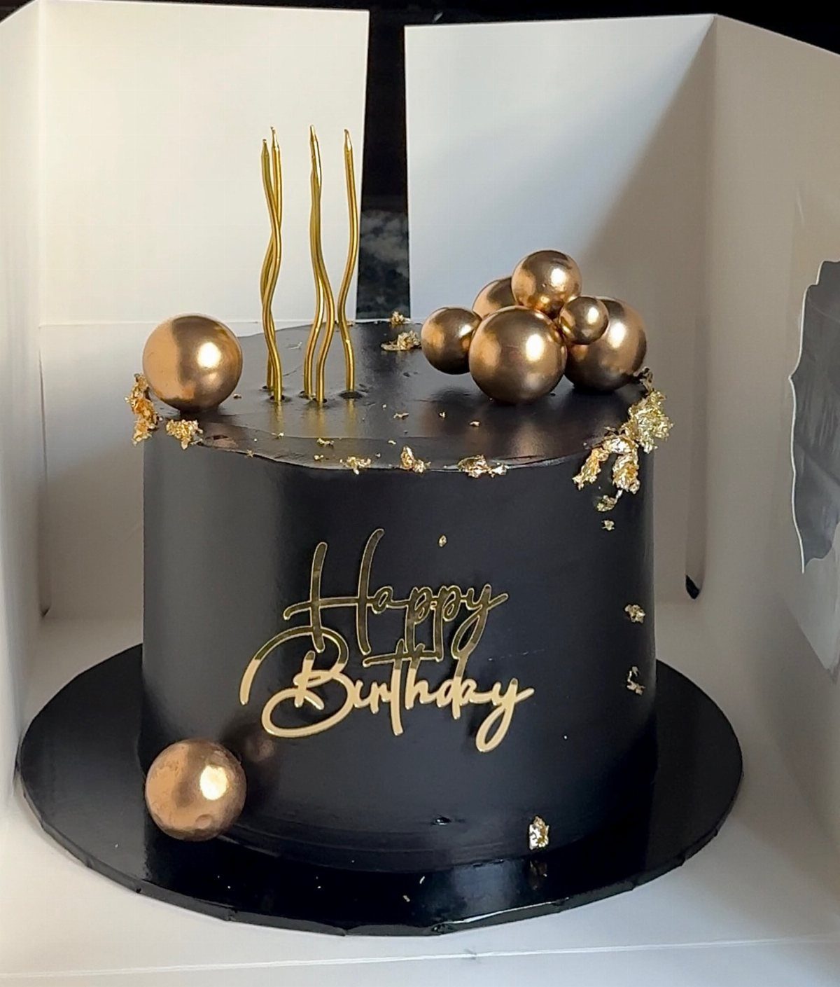 Black frosted birthday cake with gold spheres, gold leaf, and gold lettering.