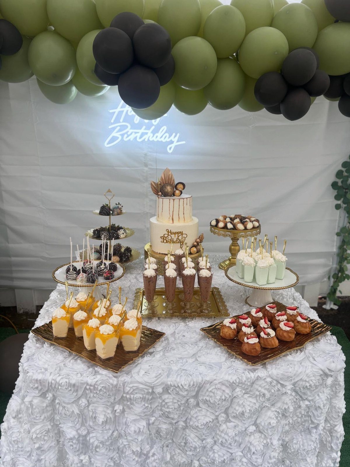 Dessert table with cake and treats under green and black balloons and neon sign