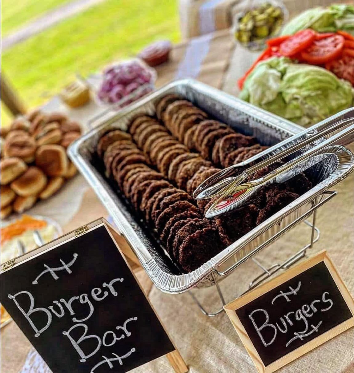 Burger bar setup with cooked patties, buns, and fresh vegetable toppings on burlap.