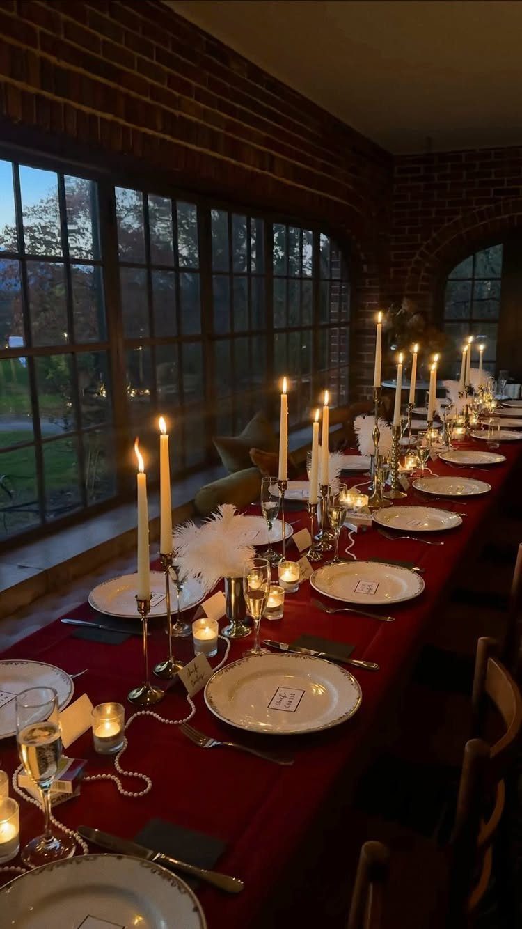 Elegant dinner table with red cloth, white candles, feathers, and pearls for a party.