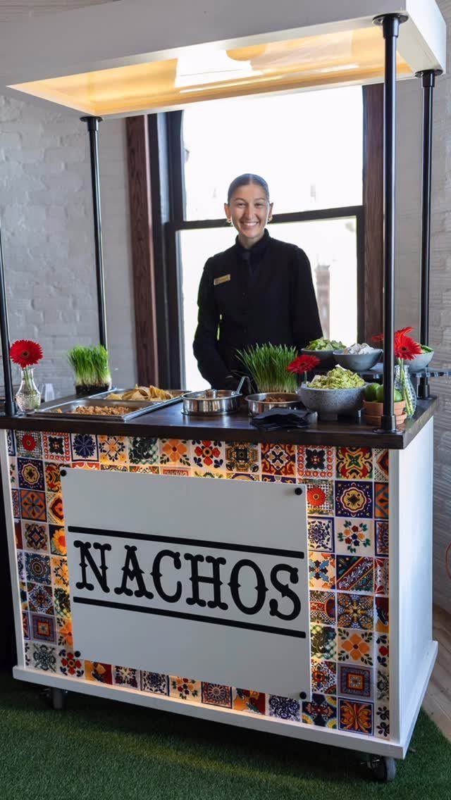 Woman standing behind a colorful nacho food cart with various toppings and chips.