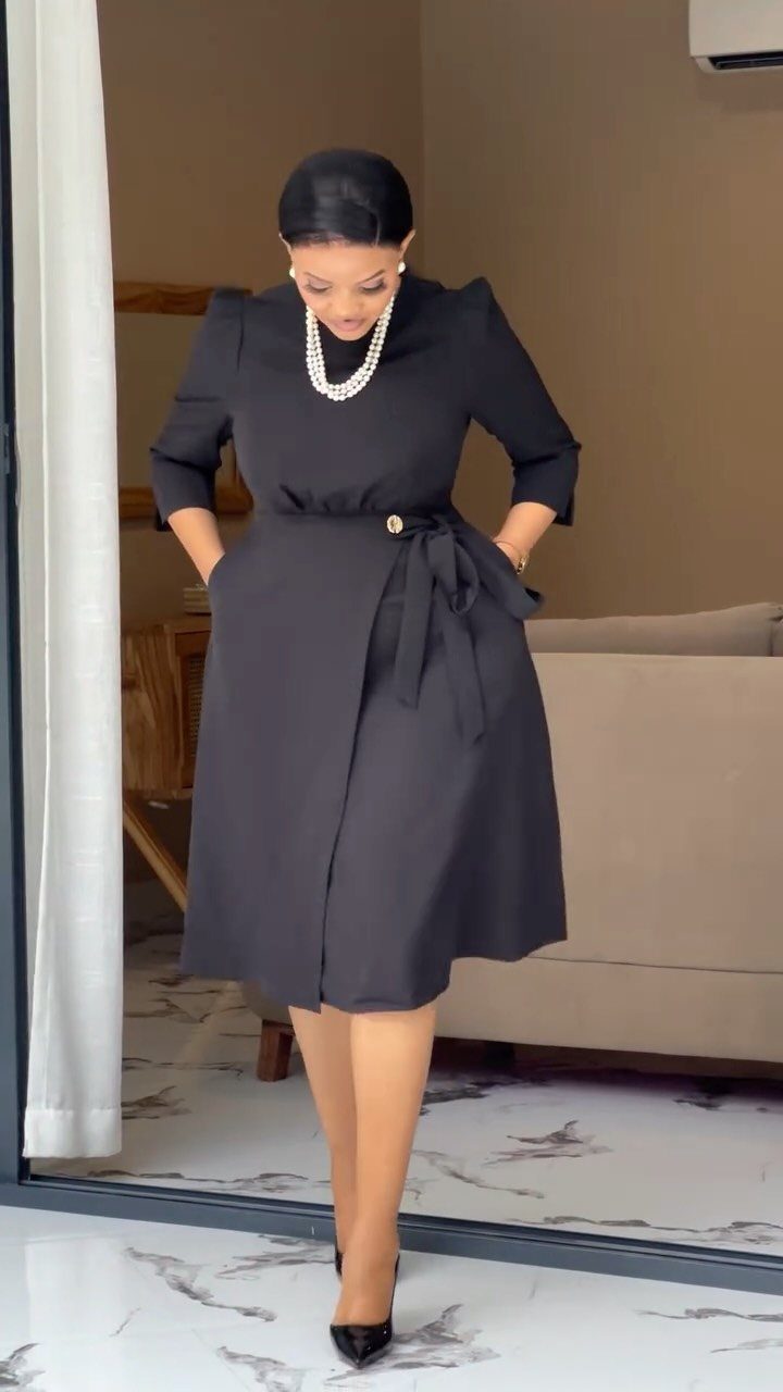 Plus-size woman in a black wrap midi dress and pearl necklace posing indoors.