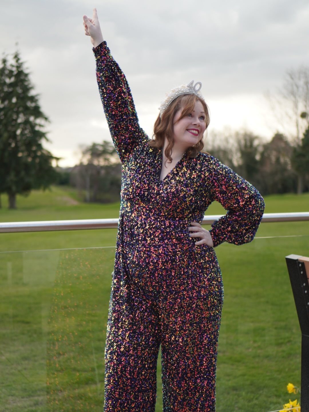 Plus-size woman wearing a multi-colored sequined jumpsuit and tiara posing outdoors.