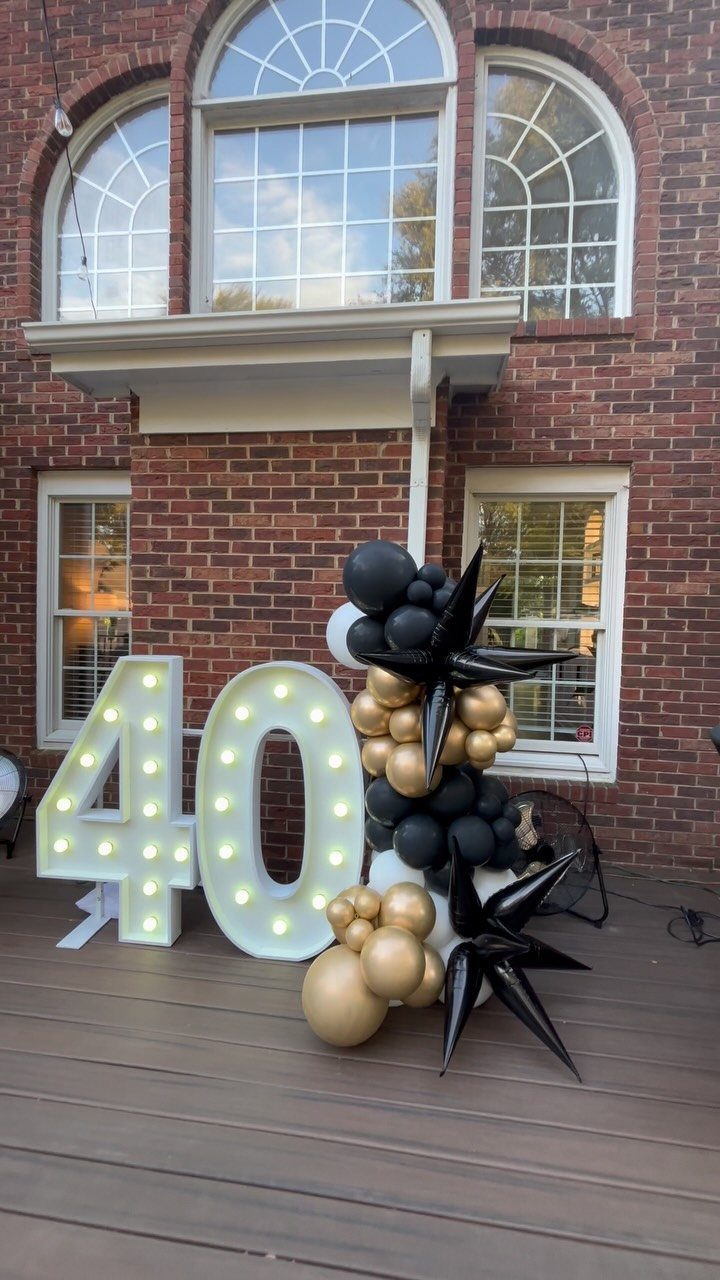 Large illuminated number 40 with black and gold balloons on a wooden deck.