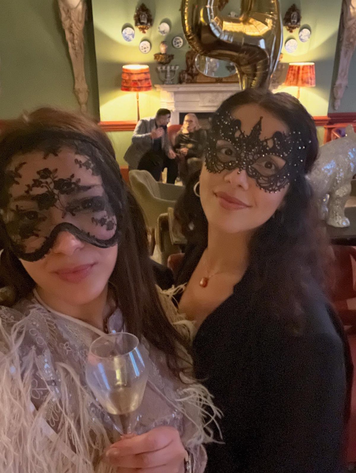 Two women wearing black lace masquerade masks and holding a glass of wine.