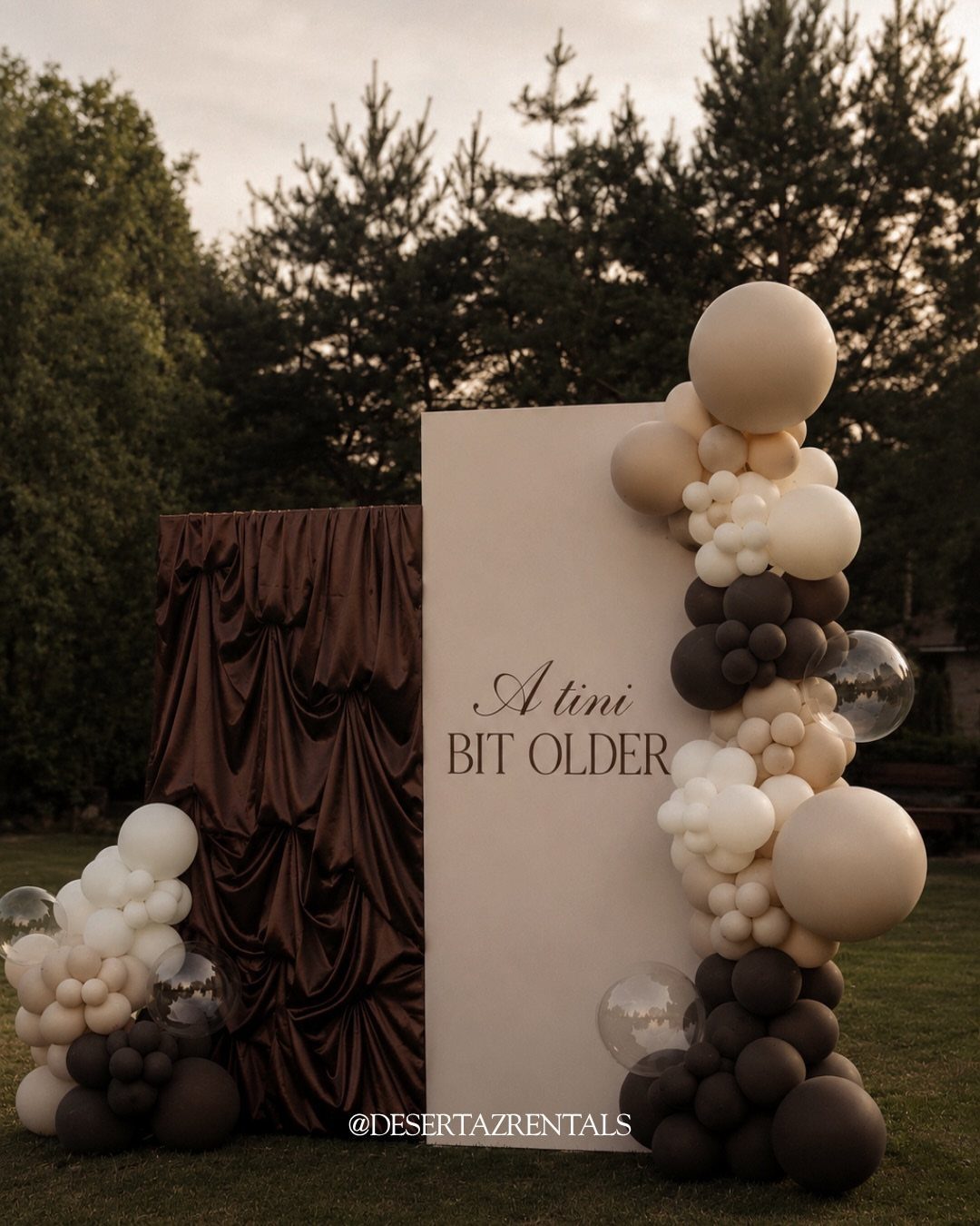 Party backdrop with cream and brown balloons and a sign saying A tini BIT OLDER