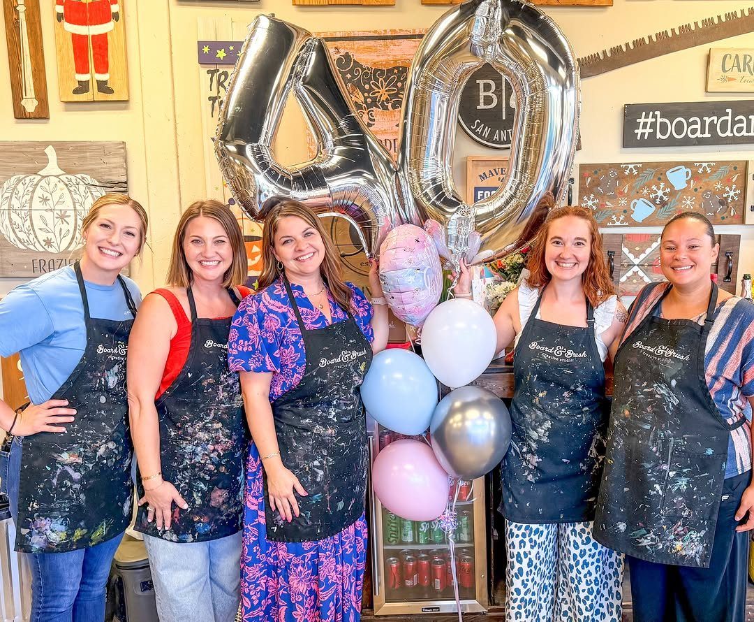 Five women in paint-splattered aprons posing with silver 40th birthday balloons in a studio.