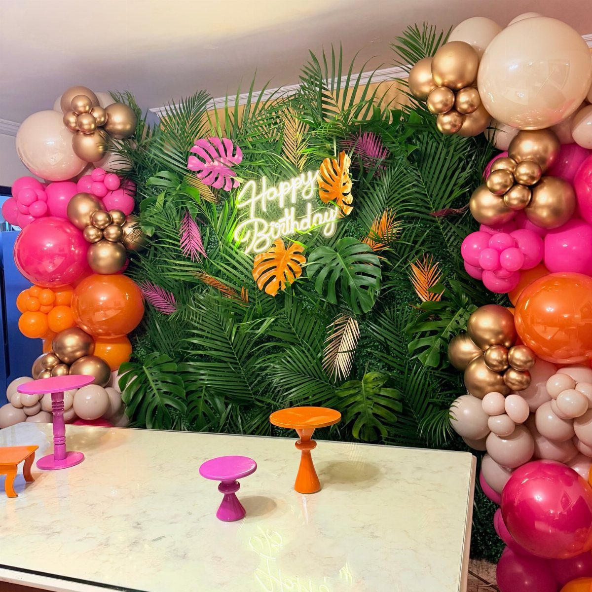 Tropical birthday party setup with palm leaf wall, neon sign, and colorful balloons.