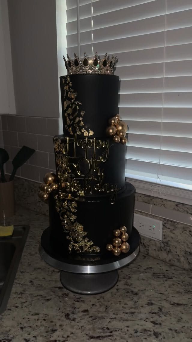 Three-tier black cake with gold accents, a 50th topper, and a crown topper.