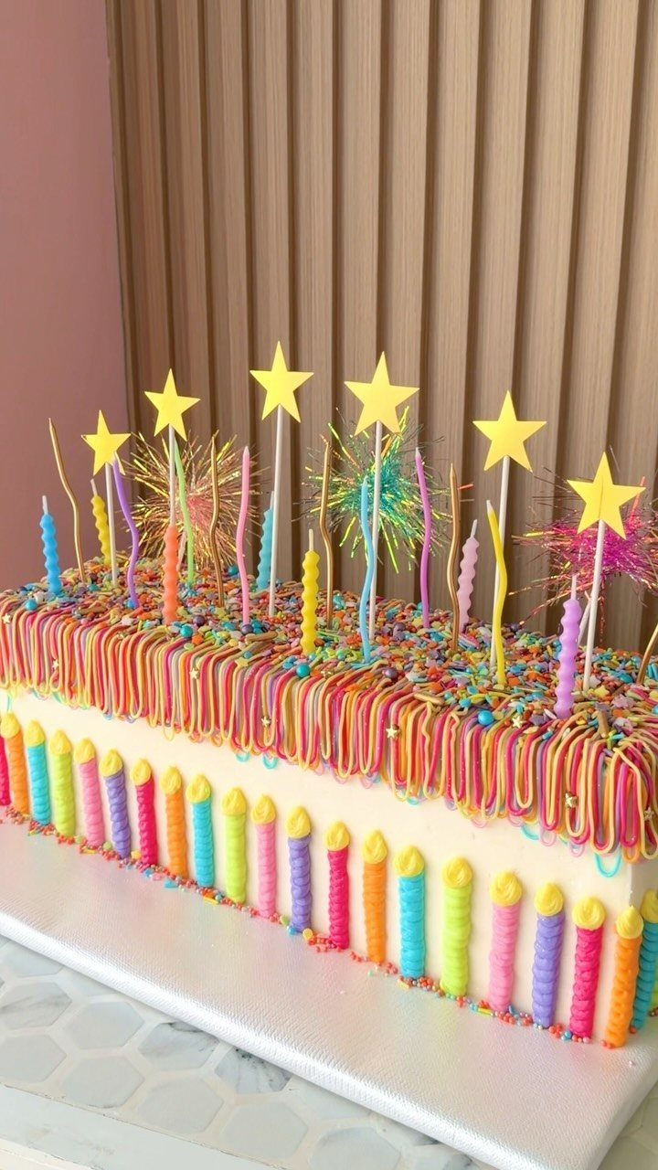 Rectangular white cake with rainbow drips, colorful candles, and yellow star toppers.