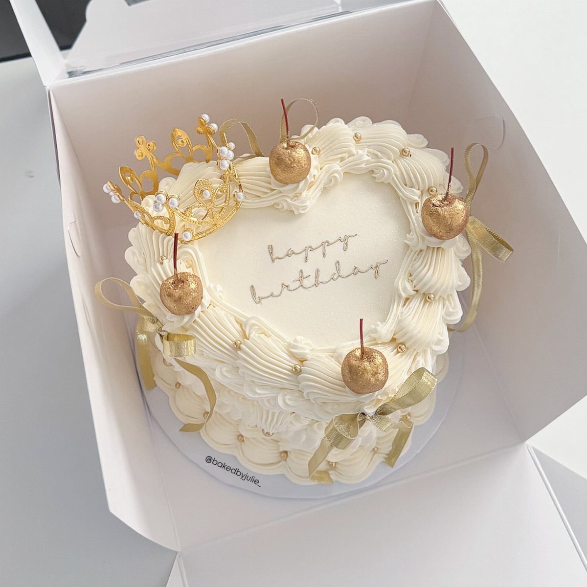 Heart-shaped white birthday cake with gold crown, gold cherries, and pearl accents.