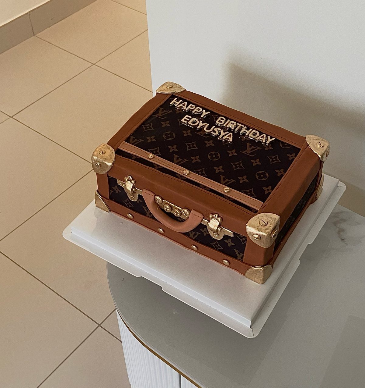 Suitcase shaped birthday cake with brown frosting, gold accents, and personalized birthday text.