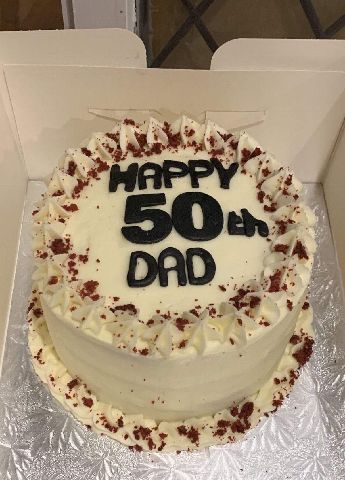 White frosted birthday cake with red crumbs and black text saying Happy 50th Dad