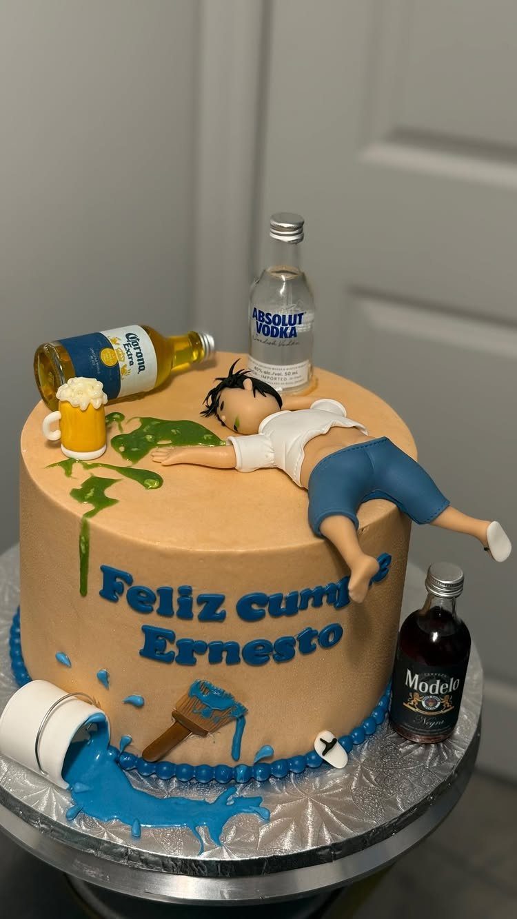 Tan birthday cake with a passed out man, alcohol bottles, and blue paint spills.